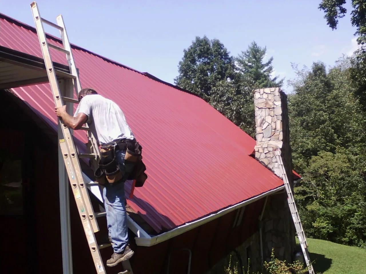 Roofer on a ladder beside a red metal roof and stone chimney — Soffit & Fascia Repair by Roofing Brothers in Whitesburg